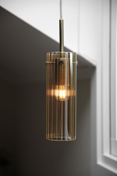 Tibia Ø7 Rav/messing vindueslampe Fra Markslöjd - Vinduslampe med cylindrisk form, riflet ravfarvet glas og elegante messingdetaljer - et varmt og dekorativt design, der giver et blødt, atmosfærisk skær i vinduet.