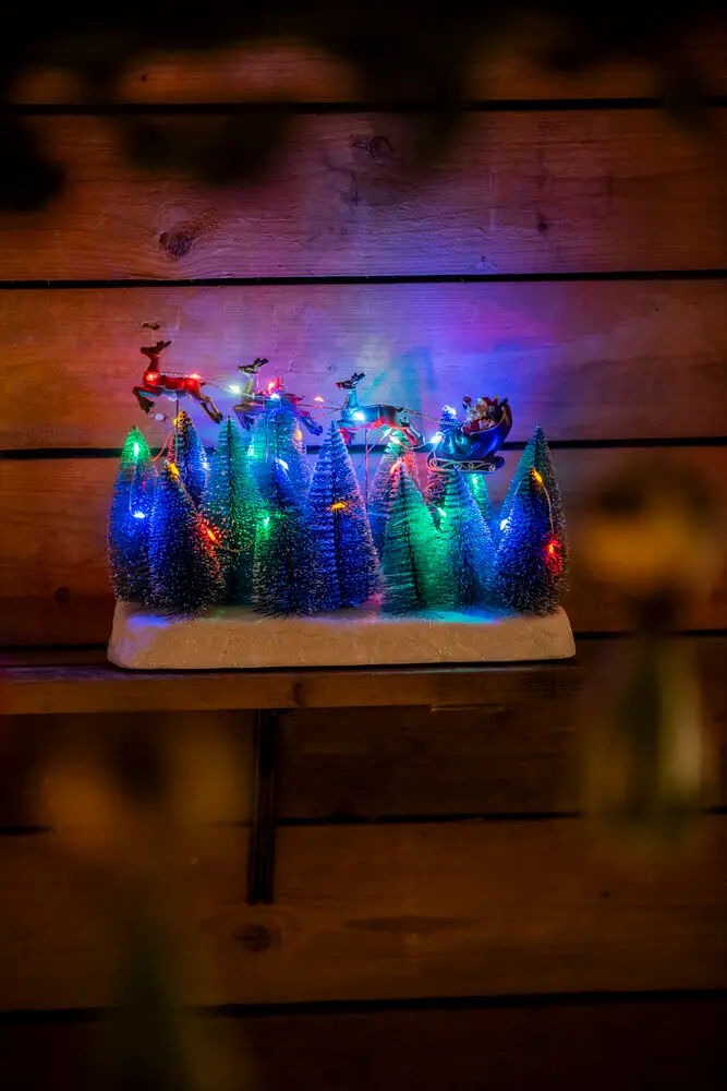 Nisse Med Slæde 22cm Flerfarvet Fra Konstsmide - Juledekoration med julemanden i en flyverdragt, der flyver over snekl&aelig;dte grantr&aelig;er.