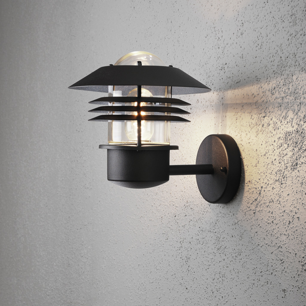 Modena Sort udendørslampe Fra Konstsmide - Med sit beskyttende tag og stilfulde design giver den et behageligt og rettet lys, ideelt til at skabe en tryg og indbydende atmosfære udendørs.