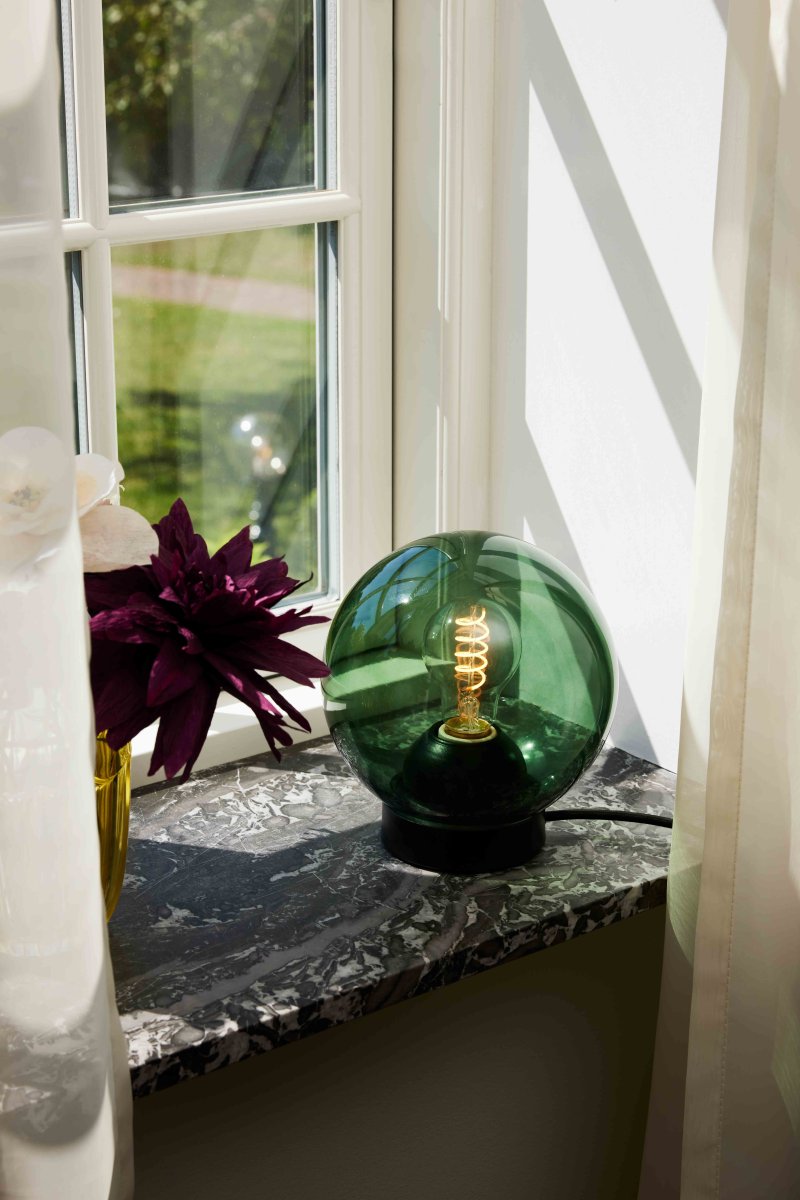 Bubbles 20cm Grøn bordlampe Fra Halo Design - Det grønne glas i kombination med de matsorte metaldetaljer bidrager til en elegance og giver en fin spredning af lys i rummet, når det er oplyst.