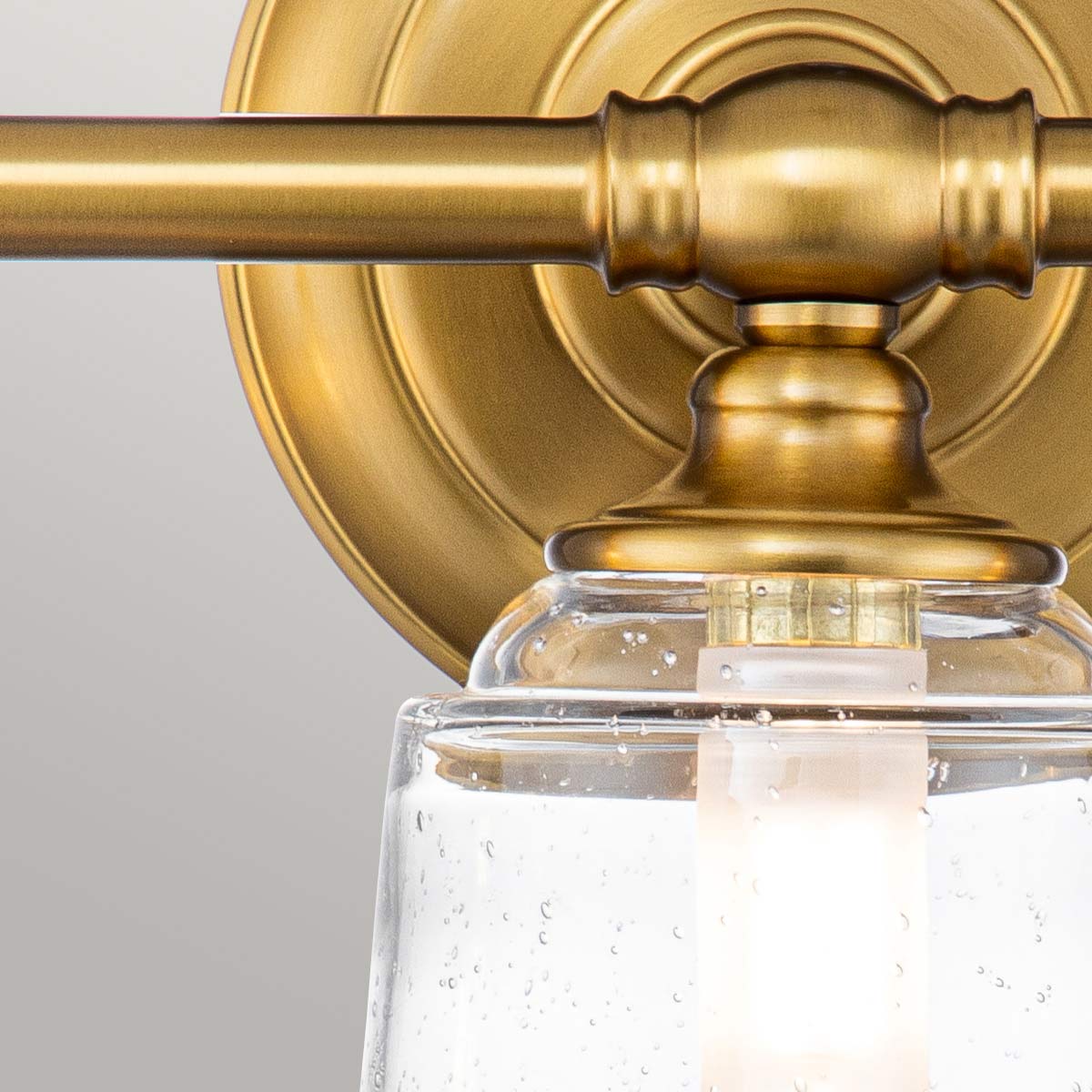 Huguenot Lake 53cm Messing badeværelseslampe Fra Elstead Lighting - De elegante cirkulære detaljer og håndskårne beslag giver en følelse af traditionelt håndværk, mens det klare, strukturerede glas skaber et blødt og indbydende lys.